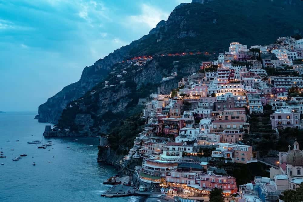 Colorful buildings cling to a steep hillside overlooking the blue ocean in Positano, Italy at dusk.