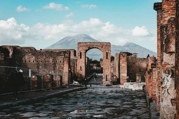 Ancient stone street in Pompeii with ruins on both sides; an arched gate frames Mount Vesuvius under a partly cloudy blue sky.