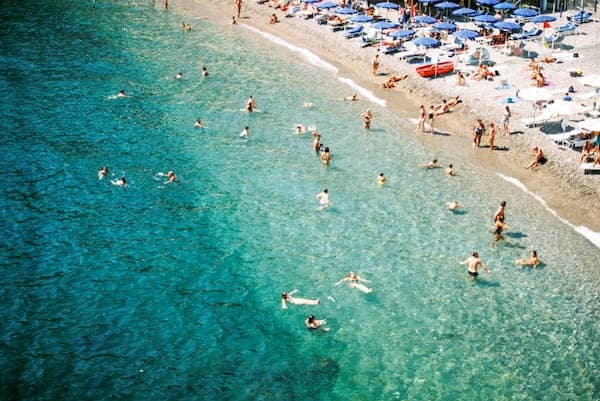 Aerial view of a beach with turquoise waters on the Amalfi Coast. People swim and relax under blue umbrellas on the sandy shore, capturing a lively, sunny atmosphere.