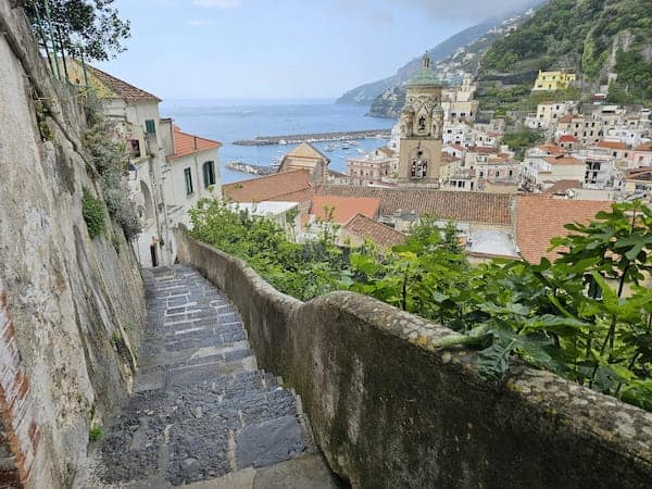 Steep stone staircase descends toward a coastal town with terracotta roofs, lush greenery, and a distant view of the sea and boats, exuding tranquillity.