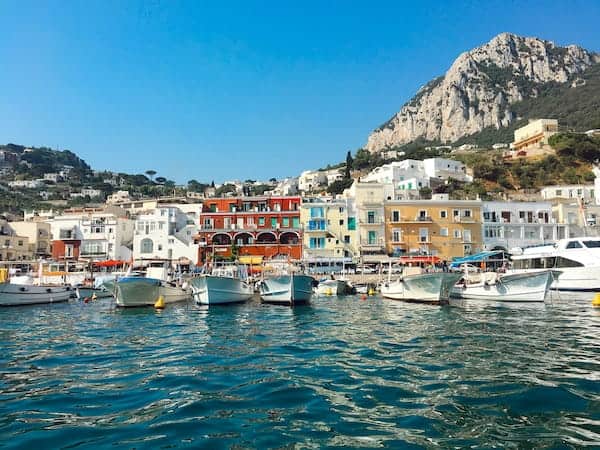 Scenic harbour in Capri, Italy, with colourful buildings and boats on turquoise water, framed by a mountainous backdrop under a clear blue sky.