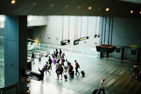 A group of travelers with luggage walking through a modern airport terminal. Overhead signs and escalators are visible. The atmosphere is busy and dynamic.